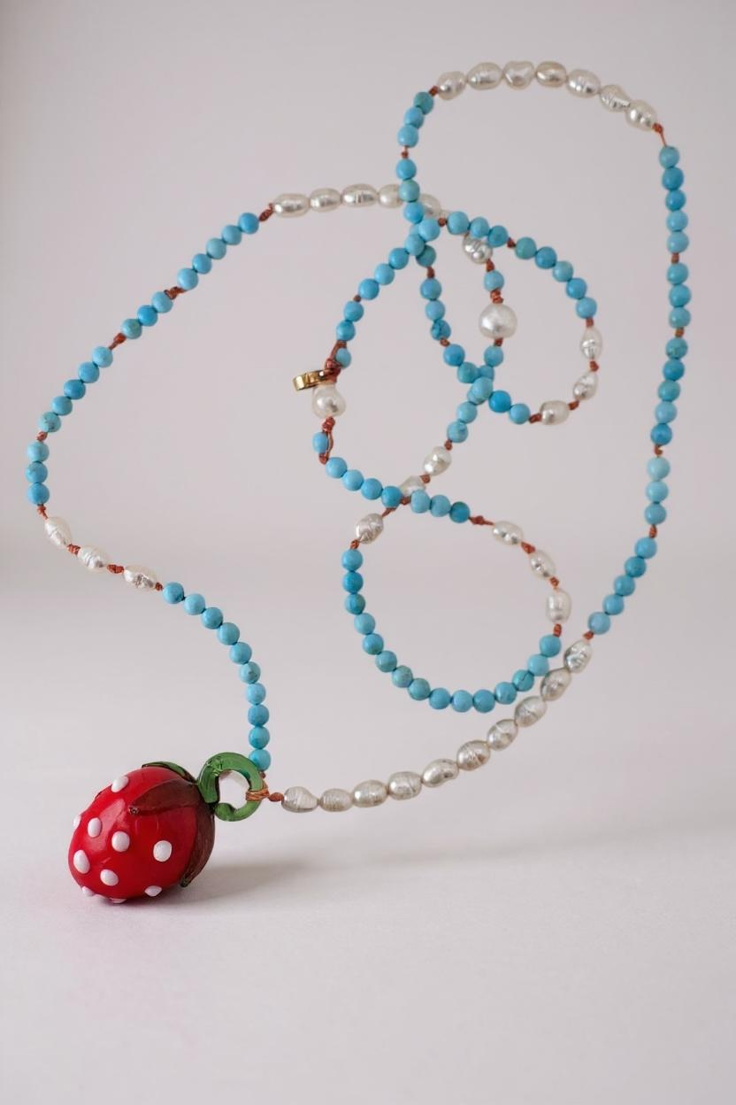Flat lay of the Strawberry Necklace on white, showing the beaded strand coiled neatly with the red glass strawberry pendant.