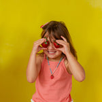 Young girl in a coral‑orange tank top holds the Strawberry Necklace pendant up to her eye against a bright yellow background