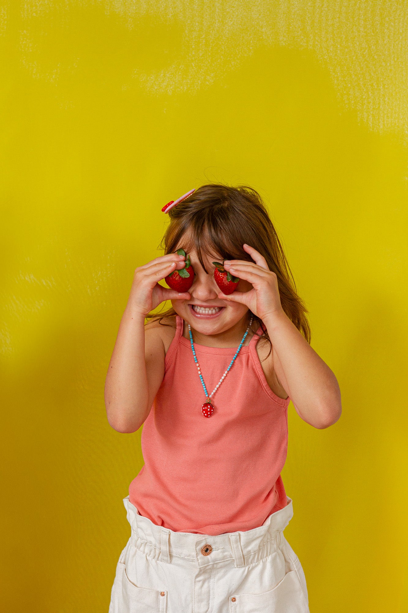 Young girl in a coral‑orange tank top holds the Strawberry Necklace pendant up to her eye against a bright yellow background