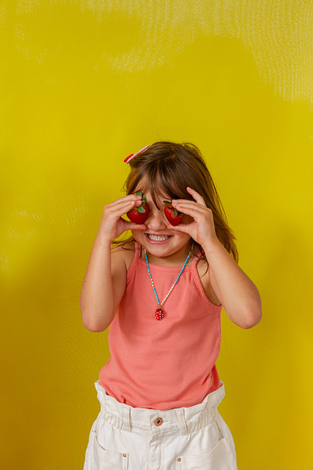 Young girl in a coral‑orange tank top holds the Strawberry Necklace pendant up to her eye against a bright yellow background