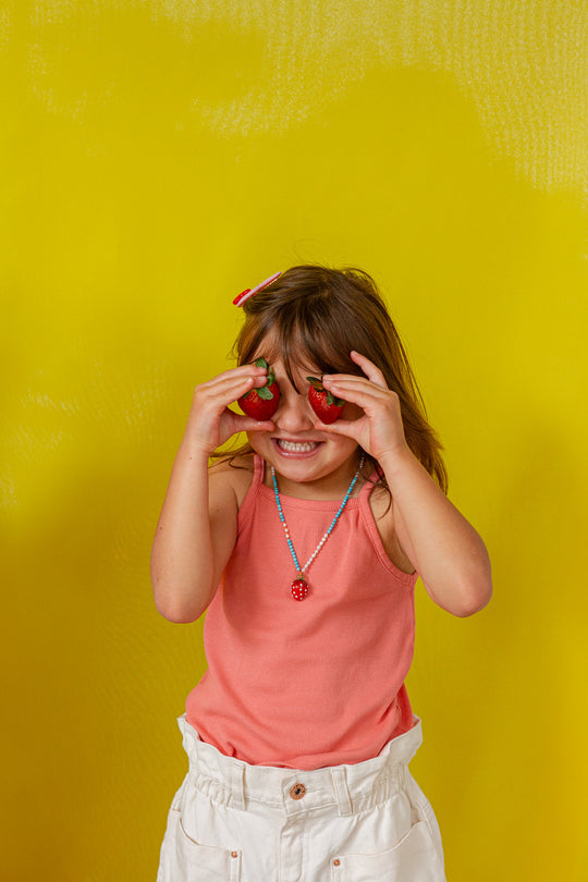 Young girl in a coral‑orange tank top holds the Strawberry Necklace pendant up to her eye against a bright yellow background