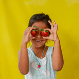 Child in a pink tank top and white shorts holds the Strawberry Necklace pendant near her face against a yellow background.