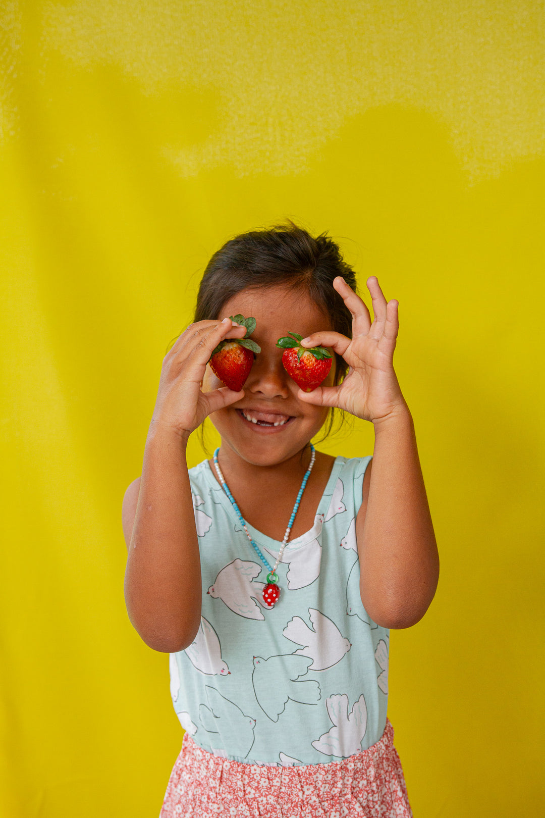 Child in a pink tank top and white shorts holds the Strawberry Necklace pendant near her face against a yellow background.