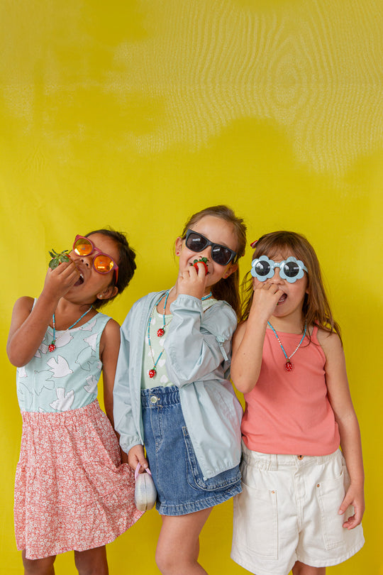 Three young girls standing together against a yellow wall, each wearing the Strawberry Necklace with the pendant hanging at varying heights.