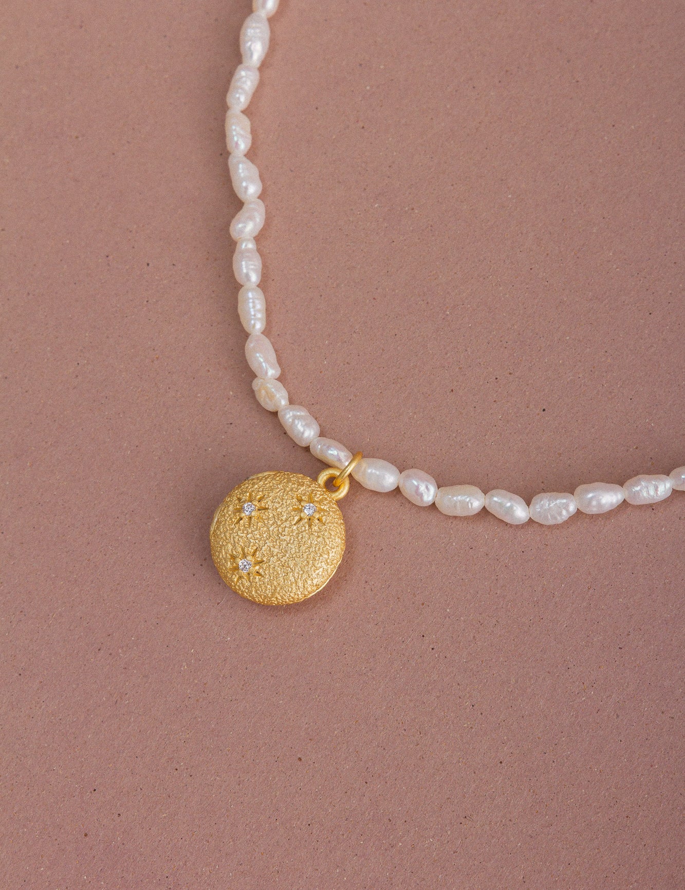 Top view of the CÂPÂ pearl locket necklace, crafted from gold-plated bronze, cultured pearls, and aquamarine-colored cubic zirconia, set against a pale pink background.
