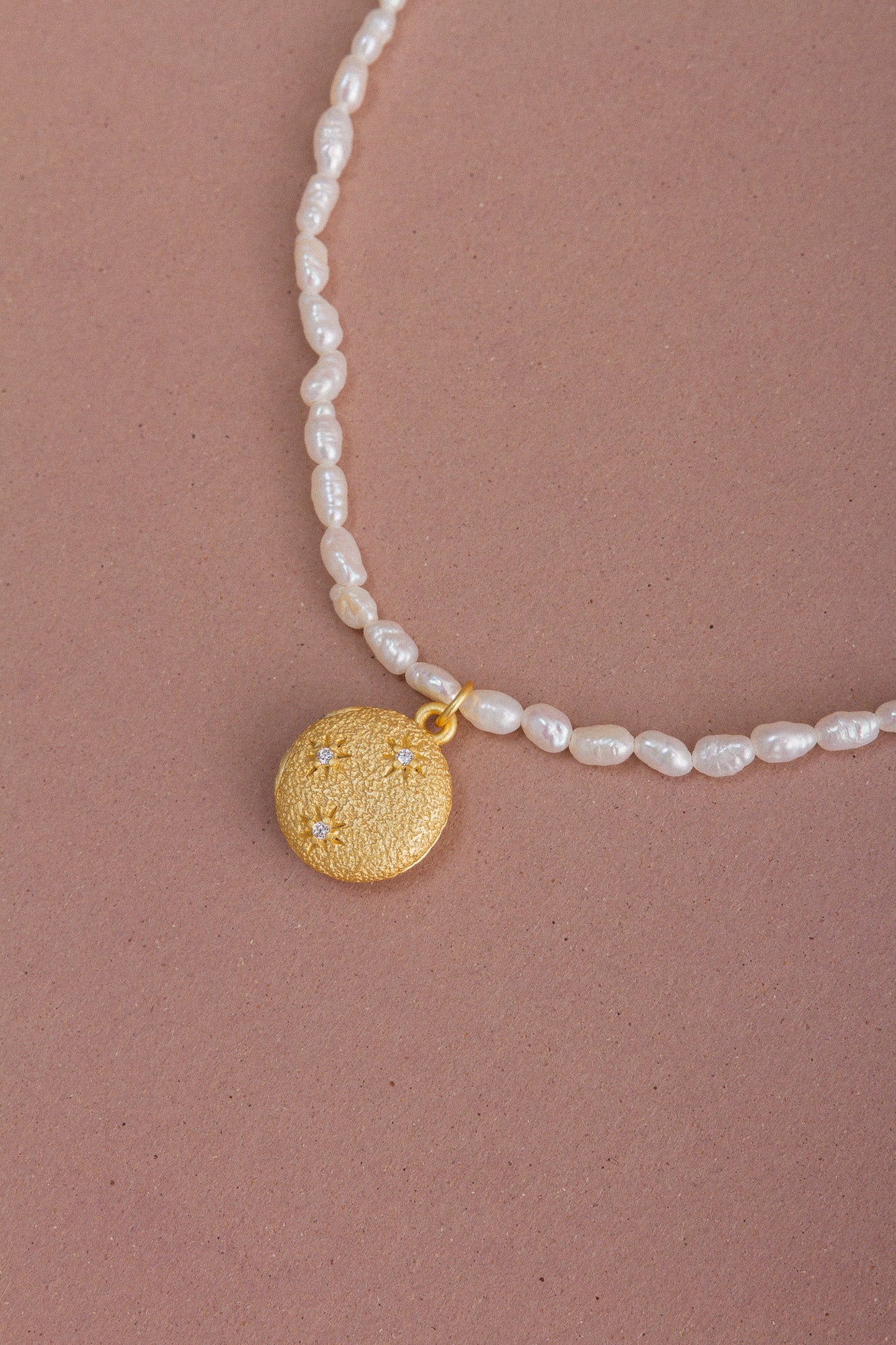 Top view of the CÂPÂ pearl locket necklace, crafted from gold-plated bronze, cultured pearls, and aquamarine-colored cubic zirconia, set against a pale pink background.
