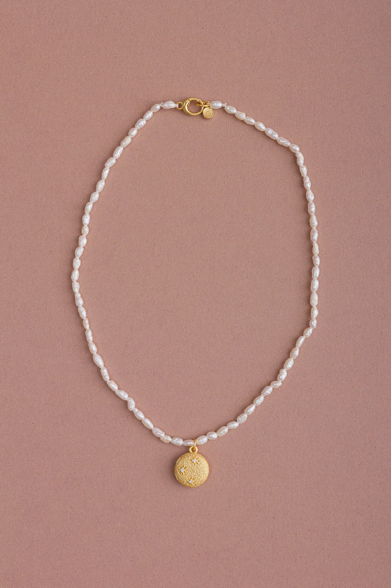 Full view of the CÂPÂ pearl locket necklace, crafted from gold-plated bronze, cultured pearls, and aquamarine-colored cubic zirconia, set against a pale pink background.