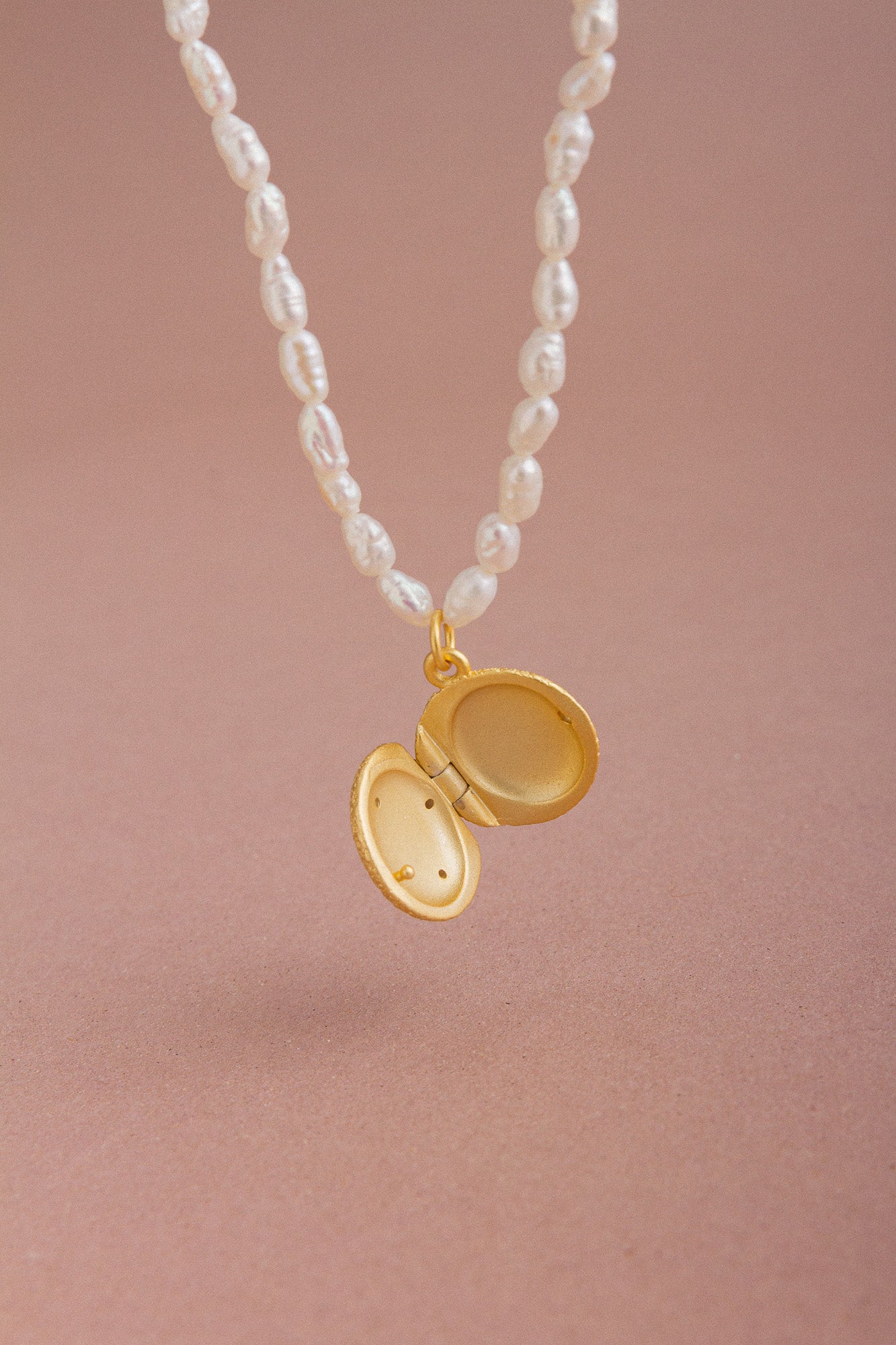 Close-up of an open CÂPÂ pearl locket necklace, crafted from gold-plated bronze, featuring cultured pearls and aquamarine-colored cubic zirconia set against a pale pink background.