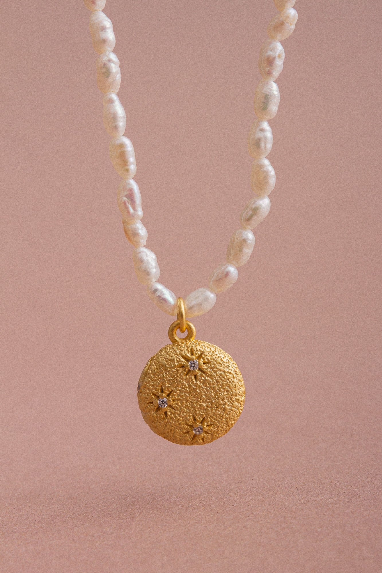 Close-up of the CÂPÂ pearl locket necklace, crafted from gold-plated bronze, featuring cultured pearls and aquamarine-colored cubic zirconia set against a pale pink background.