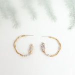 Side view of Candonga Espiga earrings in gold-plated brass with painted lavender details and glass granulation, on a blurred botanical background.