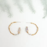 Side view of Candonga Espiga earrings in gold-plated brass with painted lavender details and glass granulation, on a blurred botanical background.