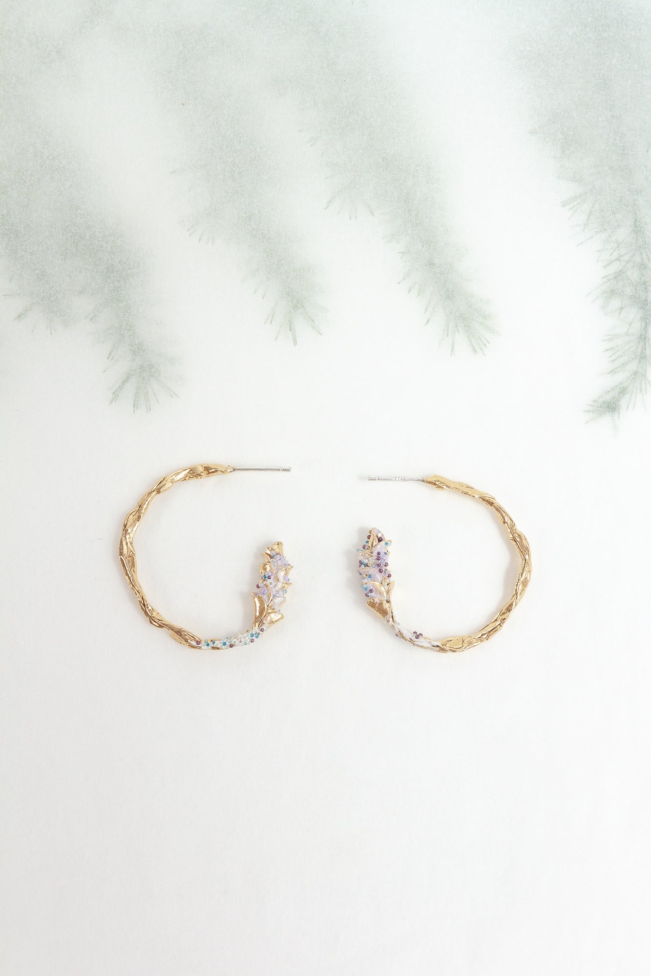 Side view of Candonga Espiga earrings in gold-plated brass with painted lavender details and glass granulation, on a blurred botanical background.