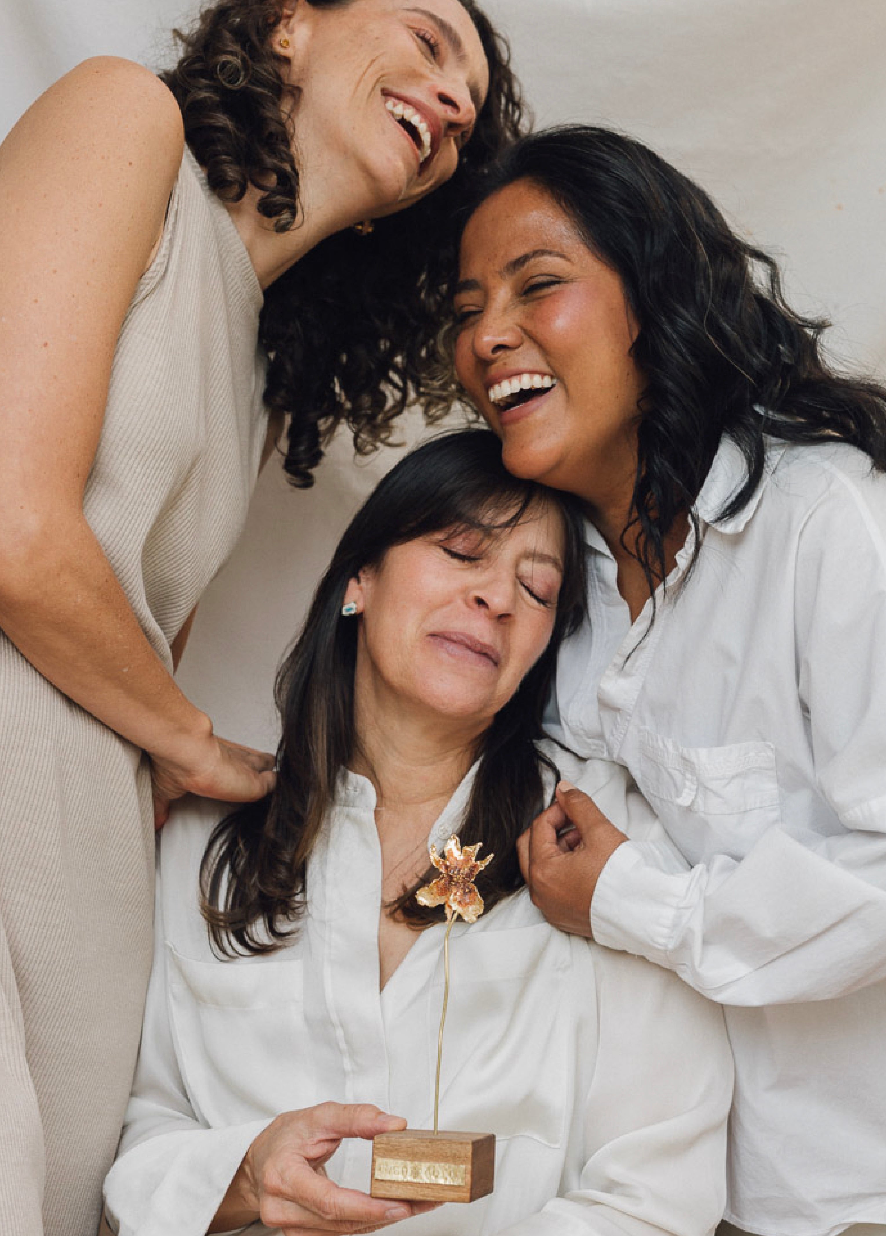 Three women sharing a joyful moment. The woman in the center, seated and smiling with her eyes closed, holds a small wooden sculpture with golden flowers. The two standing women lean toward her, laughing warmly