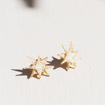 Two gold star stud earrings with white crystal detailing laid flat on a white surface, with delicate leaf-shaped shadows overhead.
