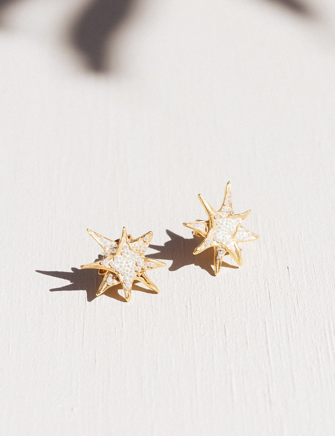 Two gold star stud earrings with white crystal detailing laid flat on a white surface, with delicate leaf-shaped shadows overhead.