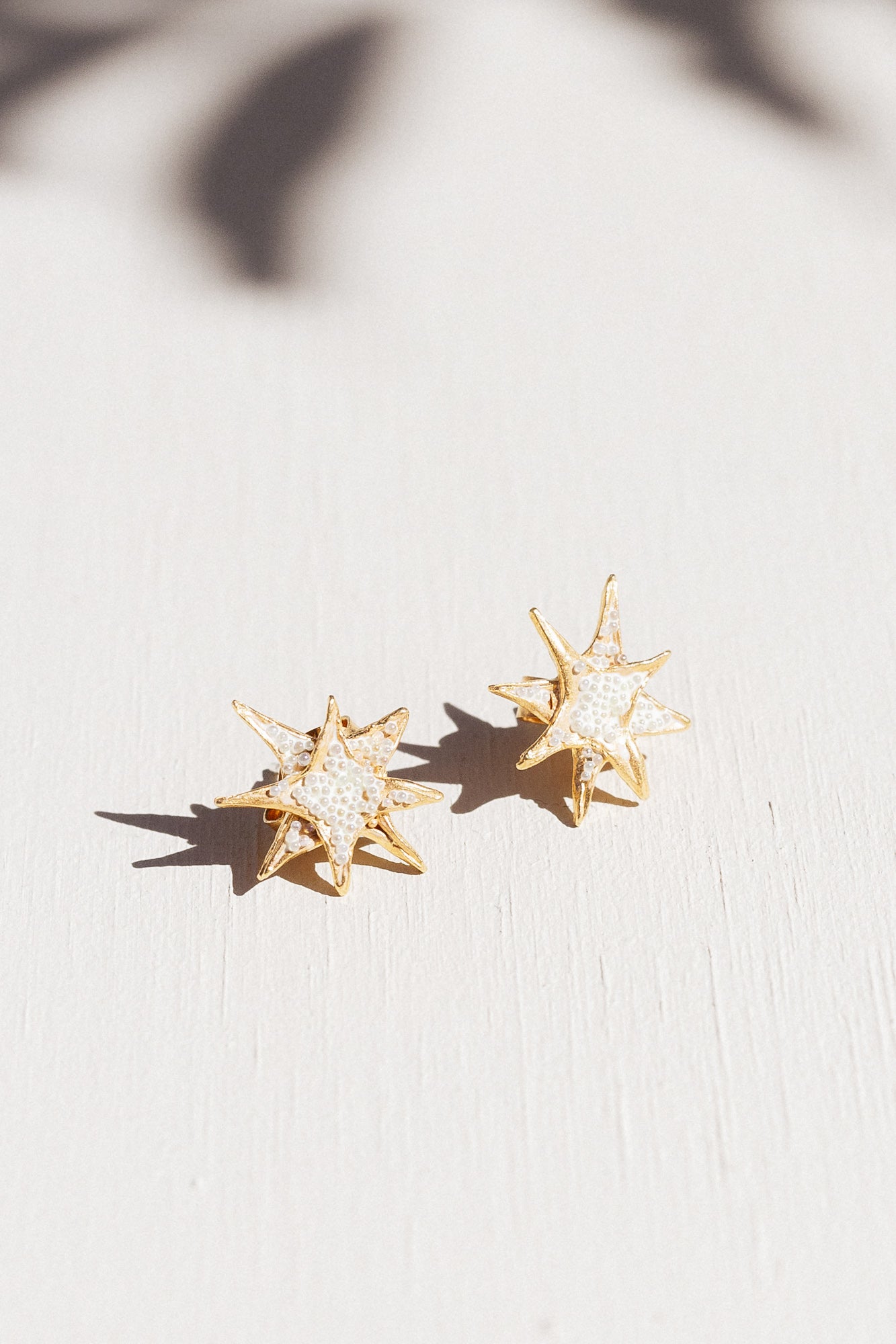 Two gold star stud earrings with white crystal detailing laid flat on a white surface, with delicate leaf-shaped shadows overhead.