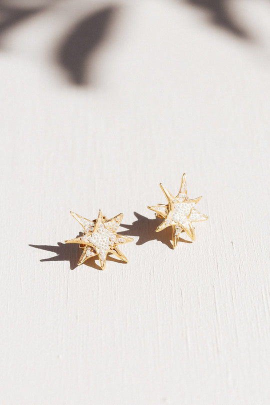 Two gold star stud earrings with white crystal detailing laid flat on a white surface, with delicate leaf-shaped shadows overhead.